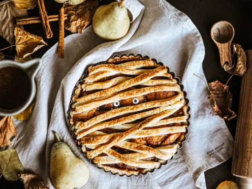 Tarte d'Halloween en forme de momie, avec des yeux globuleux et une garniture aux fruits. Autour, des feuilles d'automne, des poires et des épices.