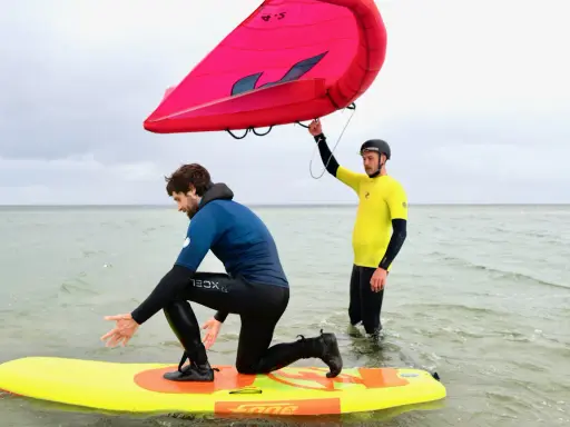Zwei Männer üben Wing Surfen im flachen Wasser. Einer kniet auf einem gelben Board, der andere hält einen roten Wing.