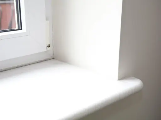 Biały parapet okienny, gotowy do wymiany. Widoczny fragment okna i ściany.