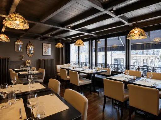 Salle à manger élégante avec des tables dressées, des luminaires Tiffany et une affiche "Les Artistes Limoges Menu" au mur.