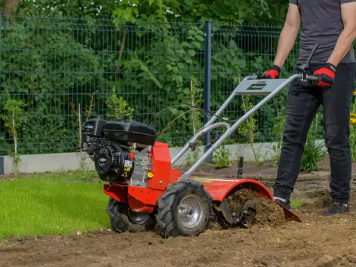 Mężczyzna w rękawiczkach obsługuje czerwoną glebogryzarkę, która spulchnia ziemię. Widać, jak działa glebogryzarka, przygotowując grunt.