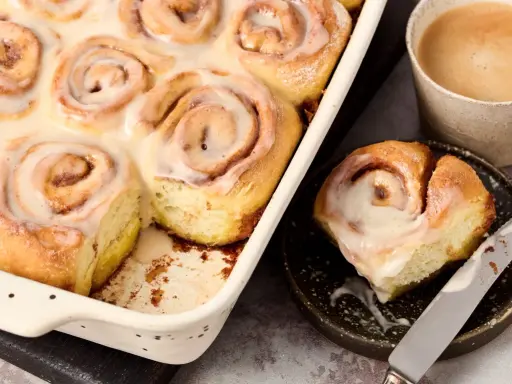 Saftige, vegane Zimtschnecken mit cremiger Glasur in einer Auflaufform, daneben eine Tasse Kaffee und eine einzelne Schnecke auf einem Teller.