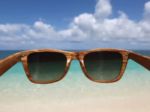 Drewniane okulary przeciwsłoneczne z filtrem uv 3 chronią oczy na plaży. W tle błękitne niebo i turkusowe morze.