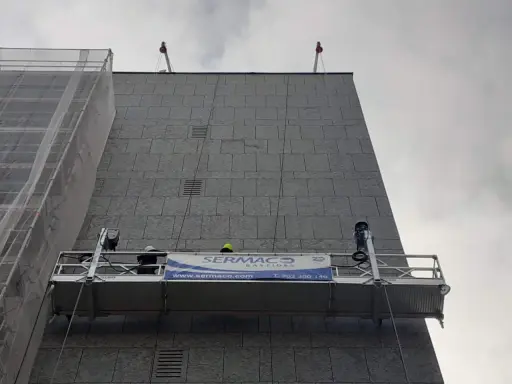 Dos trabajadores en andamios suspendidos realizan labores en la fachada de un edificio.