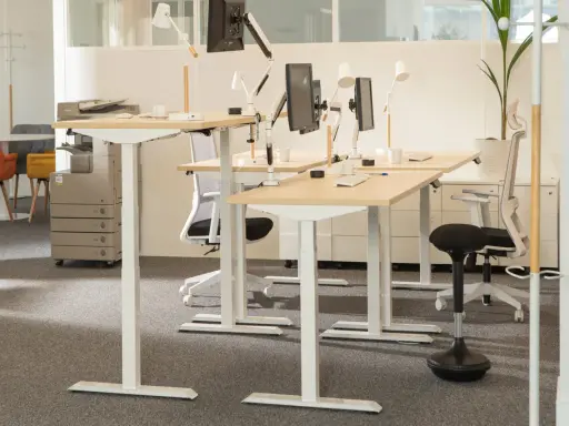 Postes de travail réglables en hauteur, favorisant l'ergonomie de bureau. Des écrans, lampes et chaises de bureau modernes sont disposés sur des bureaux en bois clair.