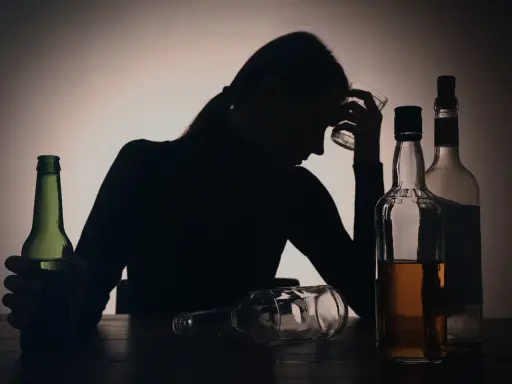 Sylwetka kobiety z kieliszkiem w dłoni, otoczona butelkami. Widoczny problem z przedawkowaniem alkoholu.