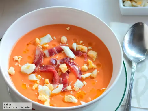 Un cuenco blanco con salmorejo cordobés, coronado con huevo duro picado y taquitos de jamón serrano.