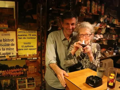 Au café du coin Le Mans, Jeanine, 73 ans, fan de rockeur, allume sa pipe. Un homme souriant se tient à ses côtés.