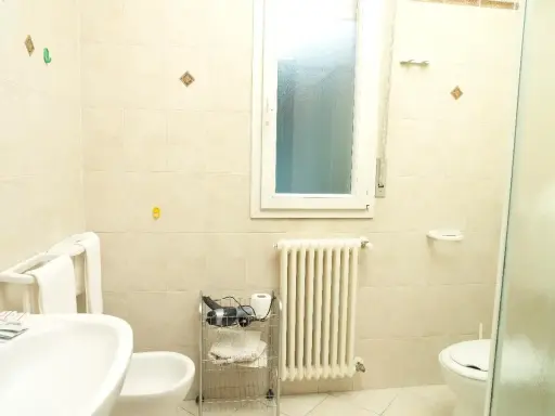 Bagno 79 Rimini: lavabo, bidet, WC, scaldasalviette e finestra. Asciugacapelli e rotolo di carta igienica su carrello.