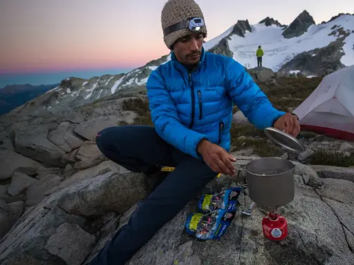 Vandrare lagar mat vid sitt msr kök i solnedgången, med tält och snöklädda berg i bakgrunden.