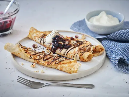 Matiga pannkakor rullade och toppade med chokladsås, blåbär och grädde. Serveras med en skål bärkompott och en blå rutig handduk.