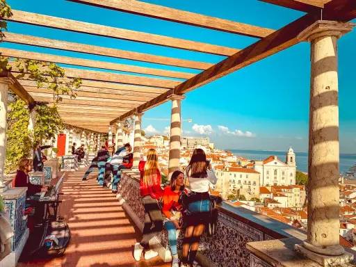 Blick vom Miradouro Lissabon über die Stadt und das Meer. Menschen genießen die Aussicht unter einer hölzernen Pergola.