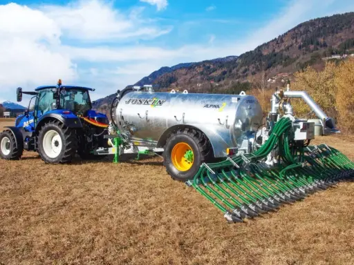 Niebieski traktor ciągnie przyczepę z cysterną do przewozu gnojowicy, wyposażoną w rozdzielacz z wieloma wężami do precyzyjnego nawożenia pól.