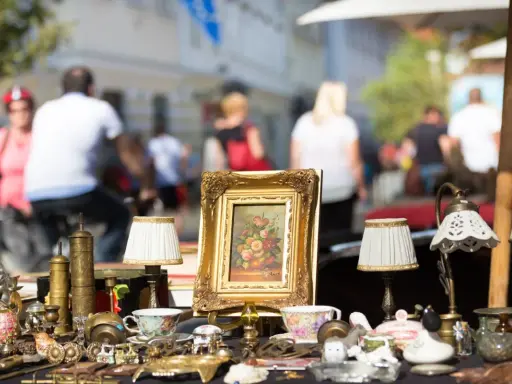 Antikviteter och kuriosa på ett bord, kanske på en loppmarknad i Stockholm. En blomstermålning i guldram, små lampor och porslin.