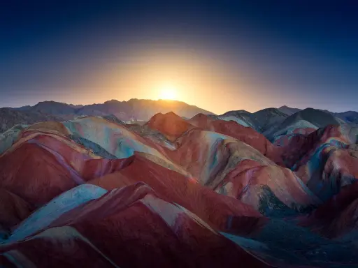 Bunte Schichten aus Rot, Gelb und Blau formen die **Regenbogenberge Chinas** unter einem tiefblauen Himmel mit aufgehender Sonne.