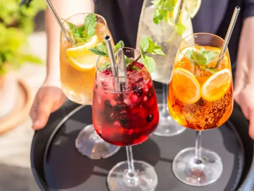 Vier erfrischende Hugo Spritz Cocktails mit Beeren, Zitrusfrüchten und Minze auf einem Tablett.