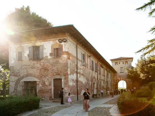 Un sole caldo illumina l'antica **abbazia di Chiaravalle**, con i suoi mattoni a vista e le persiane chiuse. Persone passeggiano nel cortile acciottolato.