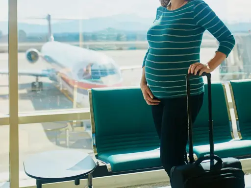 Ciężarna kobieta z walizką czeka na lotnisku. Widok na samolot przez okno. Latanie samolotem w ciąży to wyzwanie, ale podróż trwa.