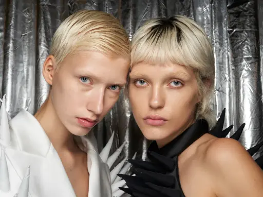 Zwei Models mit Pixie Cut feinem Haar und auffälligen Outfits vor silberner Kulisse.