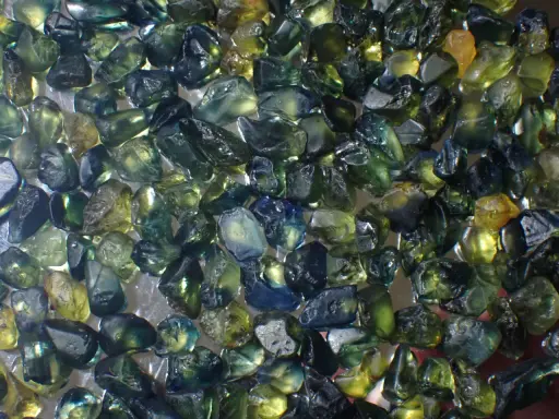 Un mucchio di zaffiri grezzi australiani, con sfumature verdi, blu e gialle. Il loro valore è legato alla rarità e alla bellezza di queste gemme naturali.