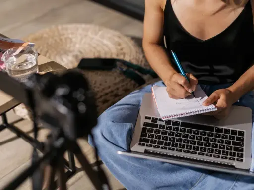 Kobieta pisze notatki w zeszycie, przygotowując materiał na artykuł. Obok leży laptop i szklanka wody.