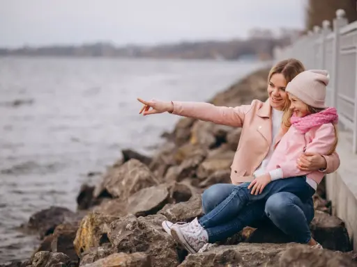 Mama z córką siedzą na kamieniach nad jeziorem. Mama wskazuje coś palcem, a dziewczynka się śmieje. To świetne gąski atrakcje dla dzieci.