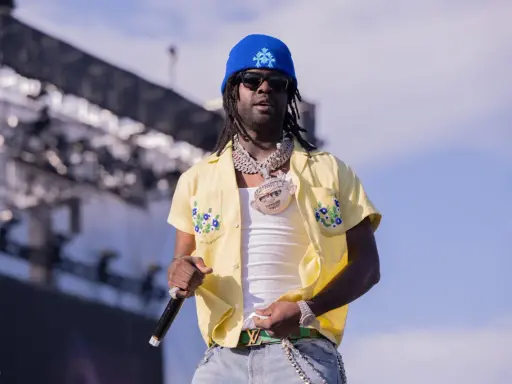 Chief Keef, w niebieskiej czapce i żółtej koszuli z kwiatami, na scenie. Jego wiek nie jest widoczny, ale styl mówi wszystko.