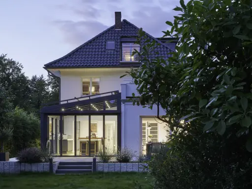 Moderner Anbau Wintergarten mit Glasfront und Beleuchtung erweitert ein Haus. Der Garten ist abends beleuchtet.