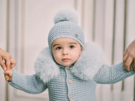 Un tierno bebé con un conjunto de punto azul claro, gorro y abrigo con cuello de pelo. Sus manos son sujetadas por adultos, como si estuviera aprendiendo a caminar. Este patrón de pelele para bebé es adorable.