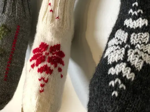 Tre stickade ärmar med mönster i maskstygn. En grå med gröna träd, en vit med röda blommor och en mörkgrå med vita snöflingor.