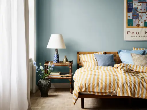 Dormitorio con paredes azul claro y ropa de cama a rayas amarillas y blancas. Los relajantes colores para dormitorios se complementan con una mesita de noche de mimbre y un jarrón con flores.