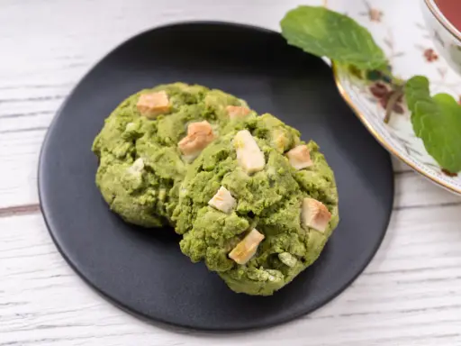 Deux cookies clairs au matcha, parsemés de morceaux de chocolat blanc, sur une assiette noire, à côté d'une tasse de thé et de feuilles de menthe.