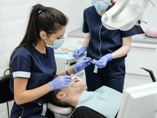 Lekarz stomatolog w masce i rękawiczkach wykonuje zabieg leczenia zębów pod narkozą. Pacjent leży na fotelu dentystycznym.
