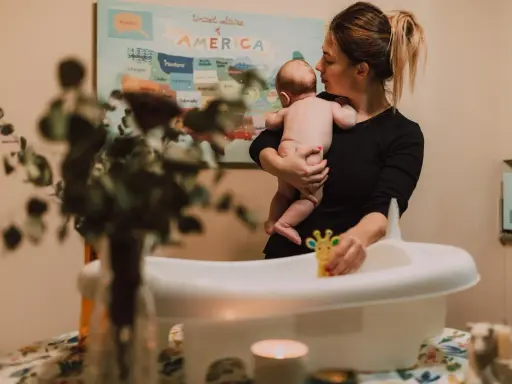 Mamma håller sitt barn redo för bad. Frågan "hur ofta bada bebis" dyker upp när man förbereder badkaret med leksaker.