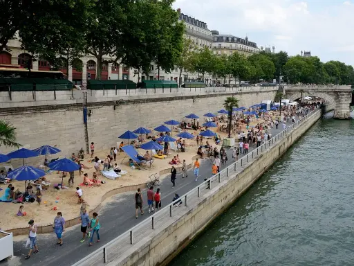 Menschen entspannen sich am **Paris strand** unter blauen Sonnenschirmen. Palmen und Gebäude säumen die Uferpromenade.