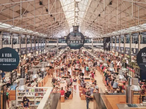 Belebter Time Out Market in Lissabon. Menschen sitzen an langen Tischen, essen und unterhalten sich. Überall gibt es Essensstände.