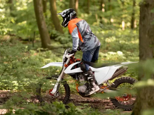 Motocyklista w kasku i pomarańczowej bluzie jedzie na białym crossie przez leśną ścieżkę.