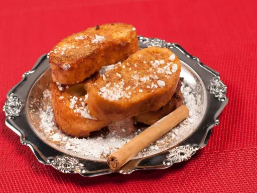 Torrijas de Semana Santa espolvoreadas con azúcar glas y canela, servidas en un plato plateado con una rama de canela.