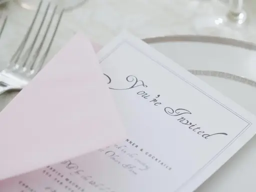 Eleganckie zaproszenie z osobą towarzyszącą, "You're Invited", na różowej kopercie, obok sztućców i talerza.