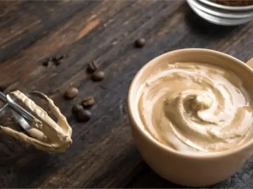 Cremige Crema al Caffè in einer Tasse, daneben ein Handmixer mit Teigresten und Kaffeebohnen auf Holz.