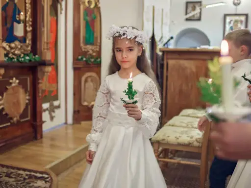 Dziewczynka w białej sukni i wianku trzyma zapaloną świecę. To ważny dzień, w którym klasa przystępuje do komunii.