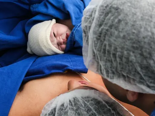 Noworodek w czepku, otulony niebieskim materiałem, leży na brzuchu mamy. Widoczna twarz dziecka i fragment twarzy mamy. To piękny moment porodu lotosowego.