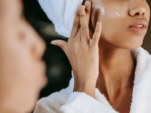 Une femme applique une crème pour l'hydratation du visage, enveloppée dans une serviette et un peignoir douillet.