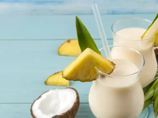 Dwa orzeźwiające koktajle Pina Colada, ozdobione kawałkami ananasa i liśćmi, na tle niebieskich desek. Idealny przepis na lato!