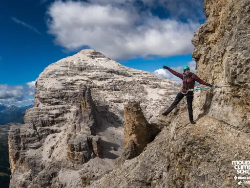 Kobieta na via ferrata dla początkujących, z rękami rozłożonymi na boki, na tle skalistych gór i błękitnego nieba.