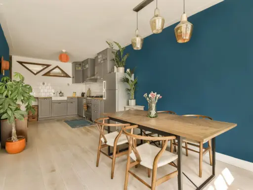 Un tavolo in legno con sedie in stile Wishbone, ideale per arredare sala da pranzo moderna. Parete blu intenso e cucina grigia completano l'ambiente.