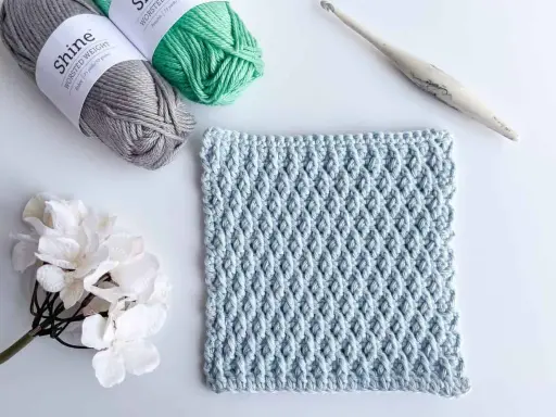 Muestra de tejido a crochet con el patrón nudo de salomon en color azul claro, junto a ovillos de lana gris y verde, una flor blanca y un ganchillo.