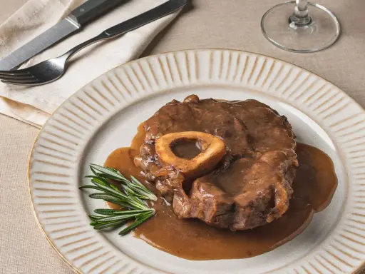 Un delicioso osso buco, tierno y jugoso, bañado en su salsa, servido en un plato con romero fresco.