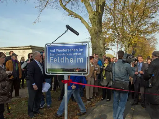 Feldheim, ein Vorreiter unter den energieautarken Gemeinden Deutschlands, wird hier mit einem Schild gefeiert. Eine Gruppe von Menschen, darunter ein Kameramann, versammelt sich.