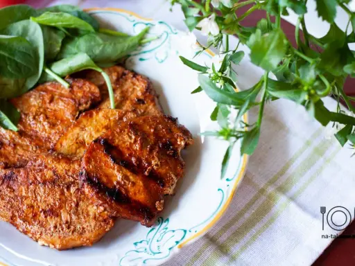Grillowane plastry schabu w aromatycznej marynacie, podane z zielonymi liśćmi na talerzu.